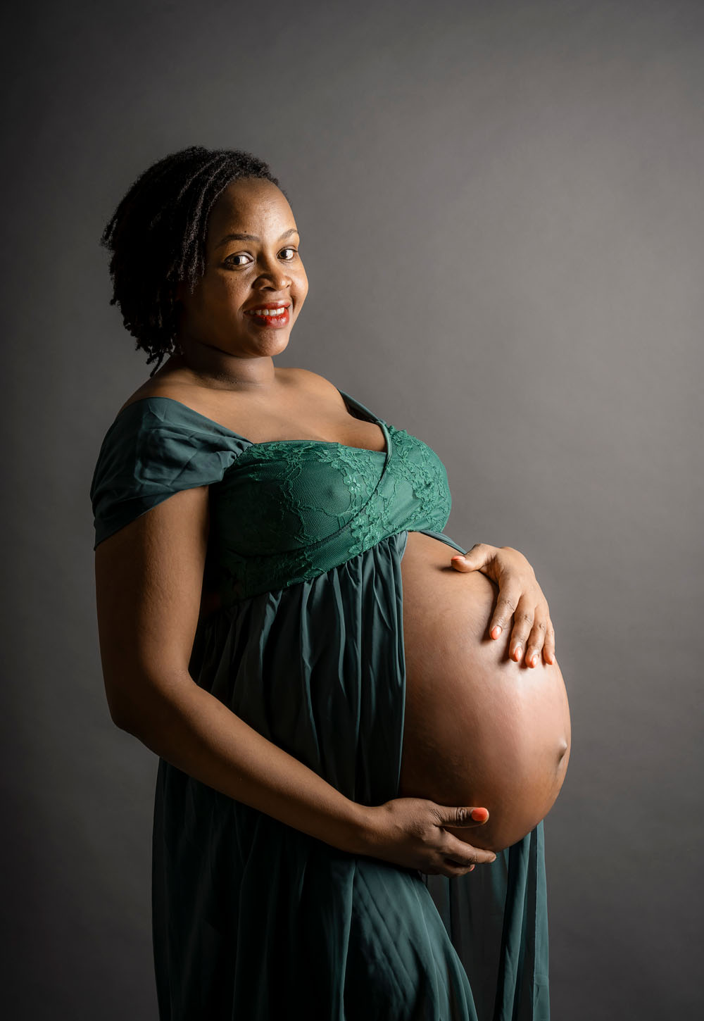 Babybauchfoto, Schwangerschaftsfoto in Farbe vor dunkelgrauen Hintergrund. Sie hält mit beiden Händen ihren Babybauch und blickt glücklich in die Kamera.