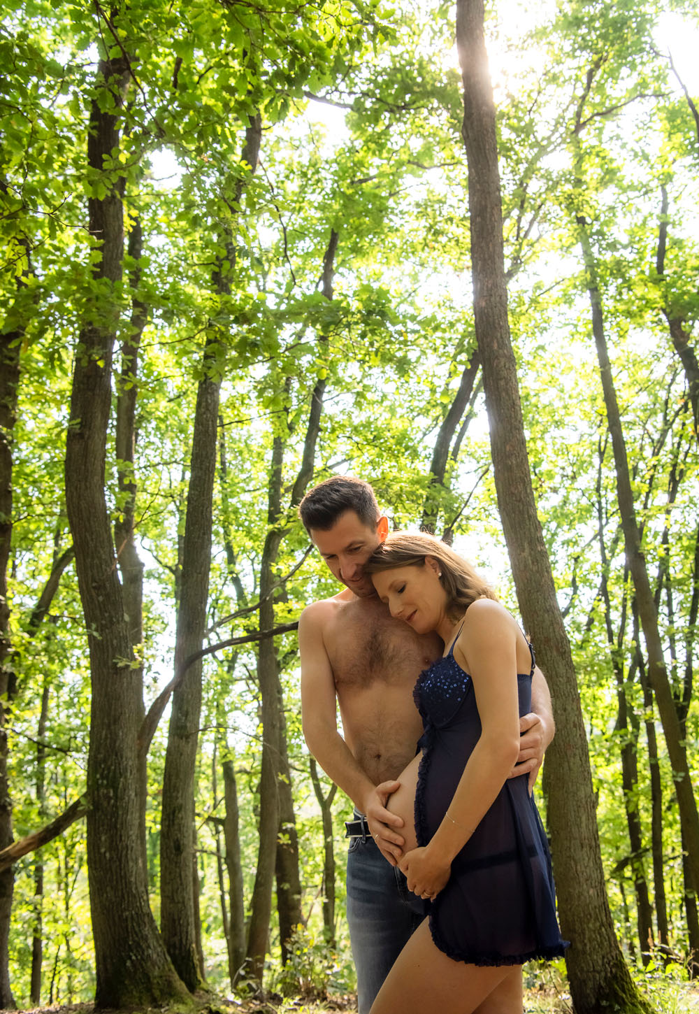 Babybauchfoto mit Partner. Beide umarmen sich innig und blicken auf den Babybauch. Dieses Schwangerschaftsfoto wurde vom Fotostudio Fischer Weinheim mit im Wald bei herrlichem Sonnenschein fotogrfaiert.