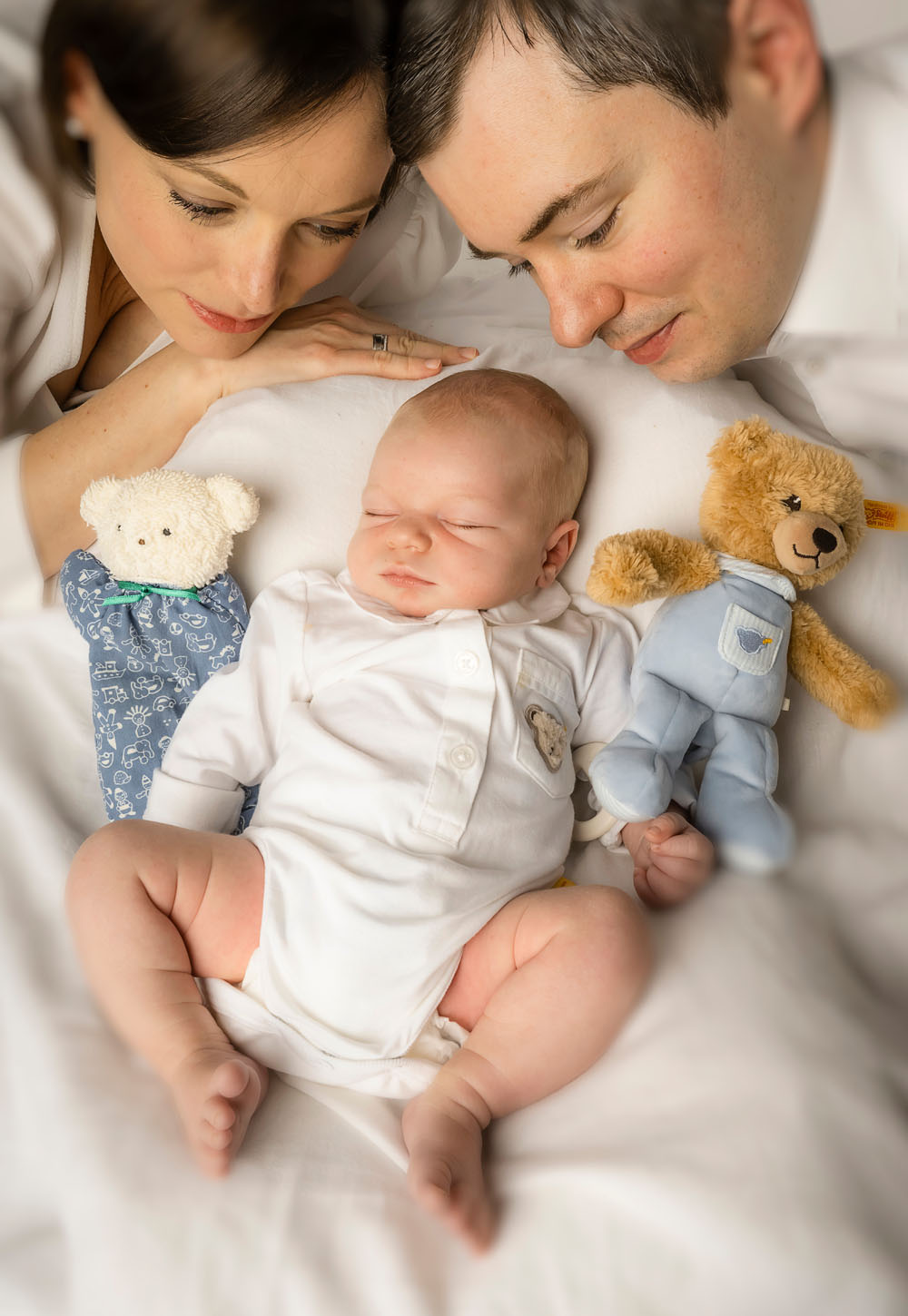 Neugeborenenfoto: Baby mit seinen Kuscheltieren und den Eltern. Eltern blicken stolz auf ihr Neugeborens. Fotografiert in Farbe bei uns im Fotostudio in Weinheim.